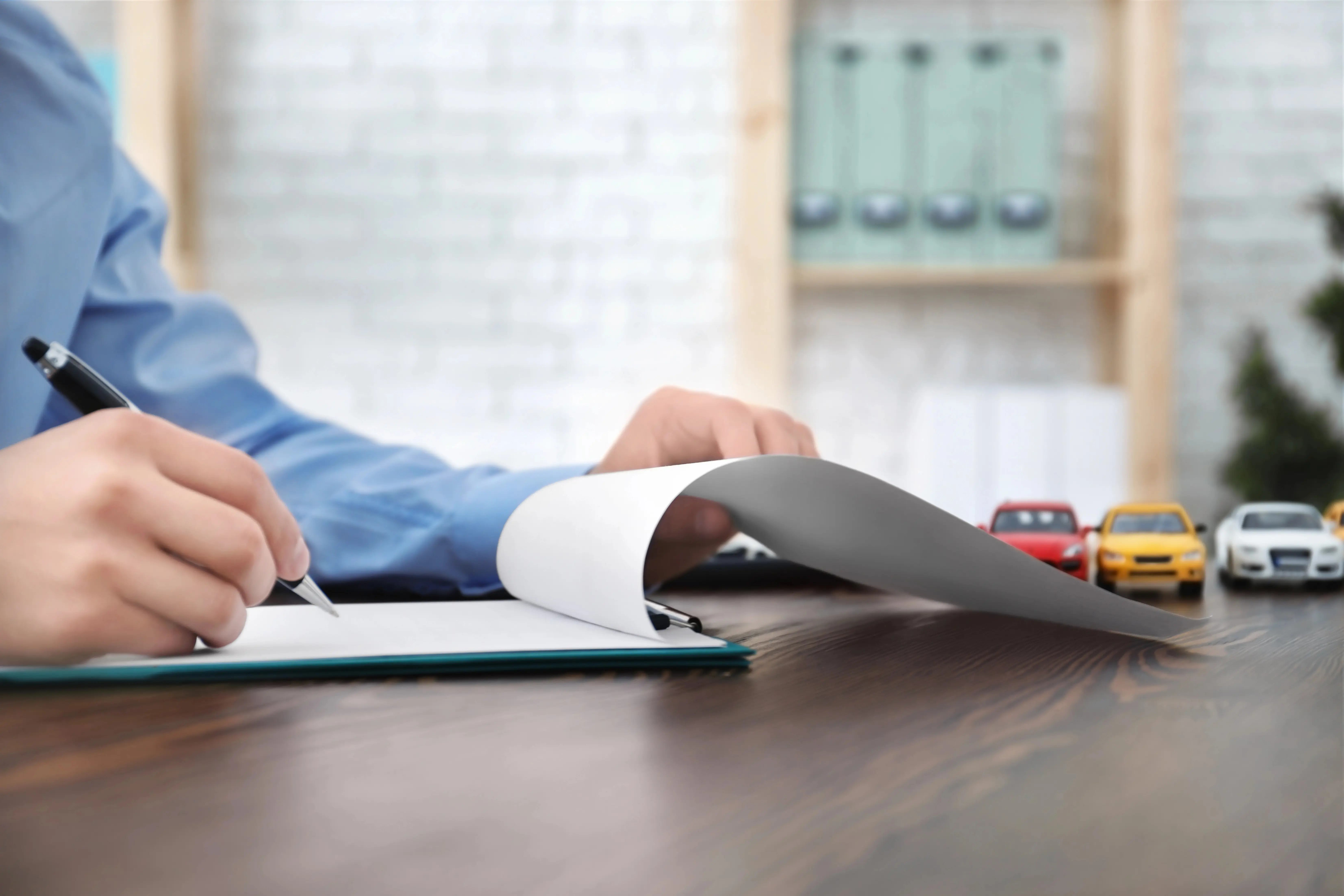 Man writing on notepad at desk with toy cars in the background.